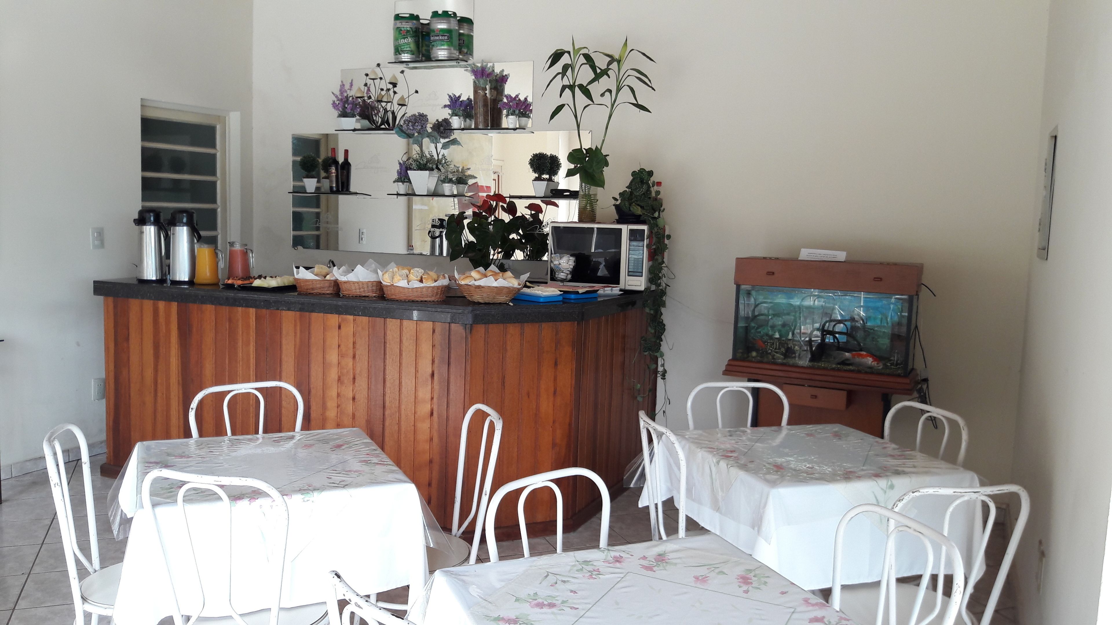 Uma área de cozinha com bancada de madeira, cadeiras brancas e uma mesa de jantar. Há diversos eletrodomésticos, um micro-ondas e um aquário sobre a bancada.