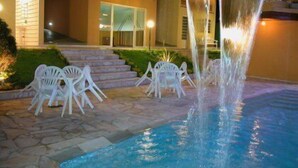 A pool area with a waterfall feature, outdoor seating, and a building with a balcony in the background. - Miragem Chales (Serra Negra)