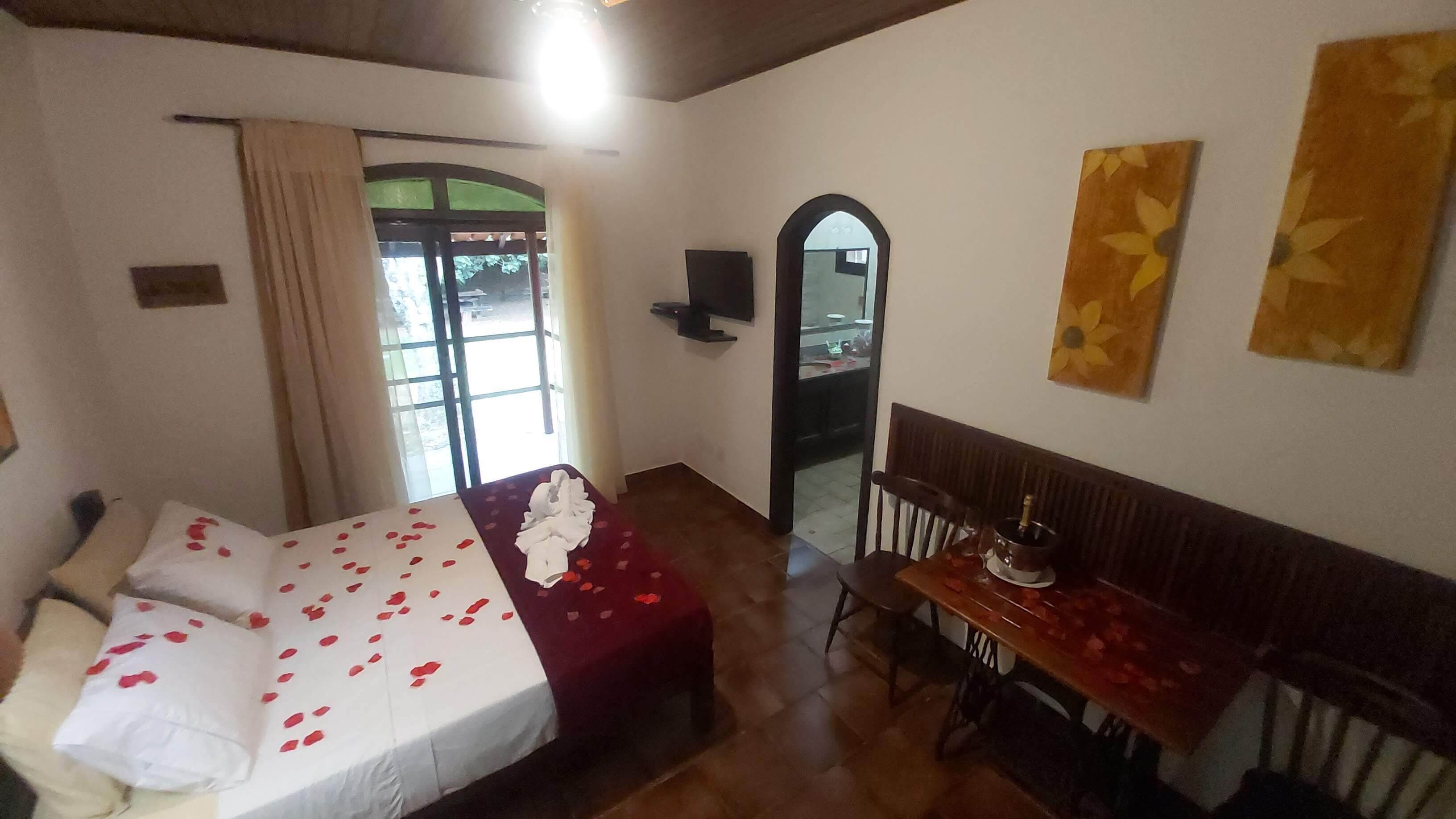 Una habitación con una cama, una mesa y un banco. Hay una ventana con cortinas, un espejo y una puerta.