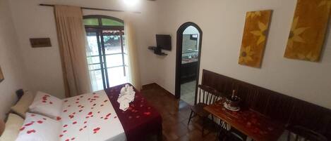 Una habitación con una cama, una mesa y un banco. Hay una ventana con cortinas, un espejo y una puerta.