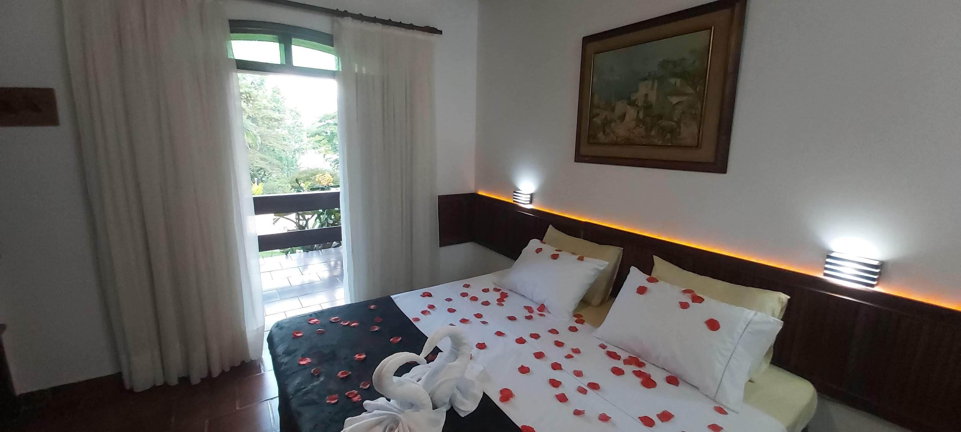 Ein Hotelzimmer mit einem Bett, das mit Rosenblättern und einer Schwan-Dekoration geschmückt ist, einem Gemälde an der Wand und einem mit Vorhängen versehenen Balkon.