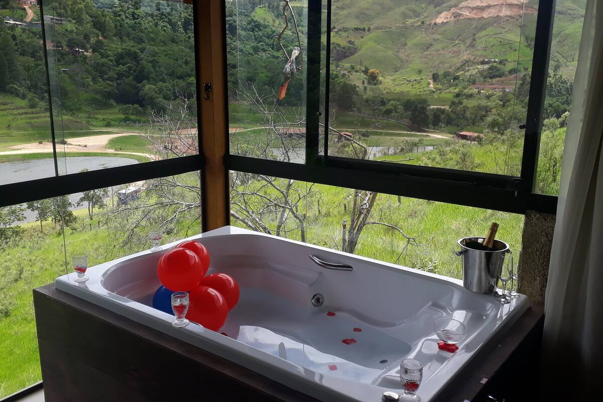 A bathroom with a bathtub, a window with a view of a river and hills, and a bottle of champagne.