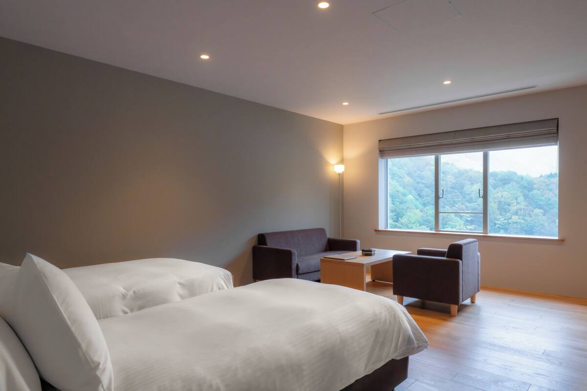 Habitación de hotel moderna con dos camas, un escritorio y una silla. Amplia ventana con vistas a árboles.