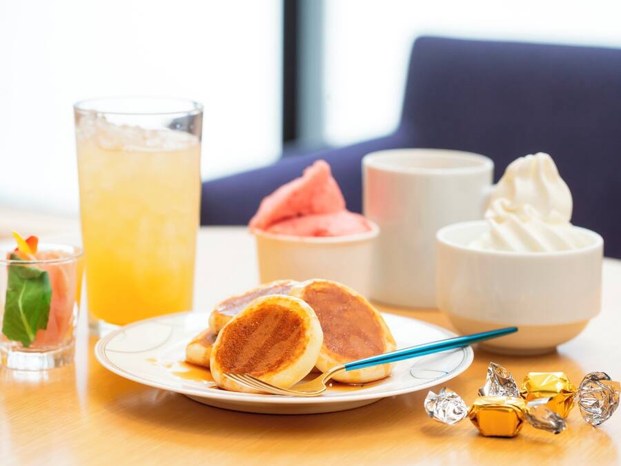 A plate of pancakes with syrup, a glass of orange juice, a bowl of whipped cream, and some wrapped chocolates on a wooden table.