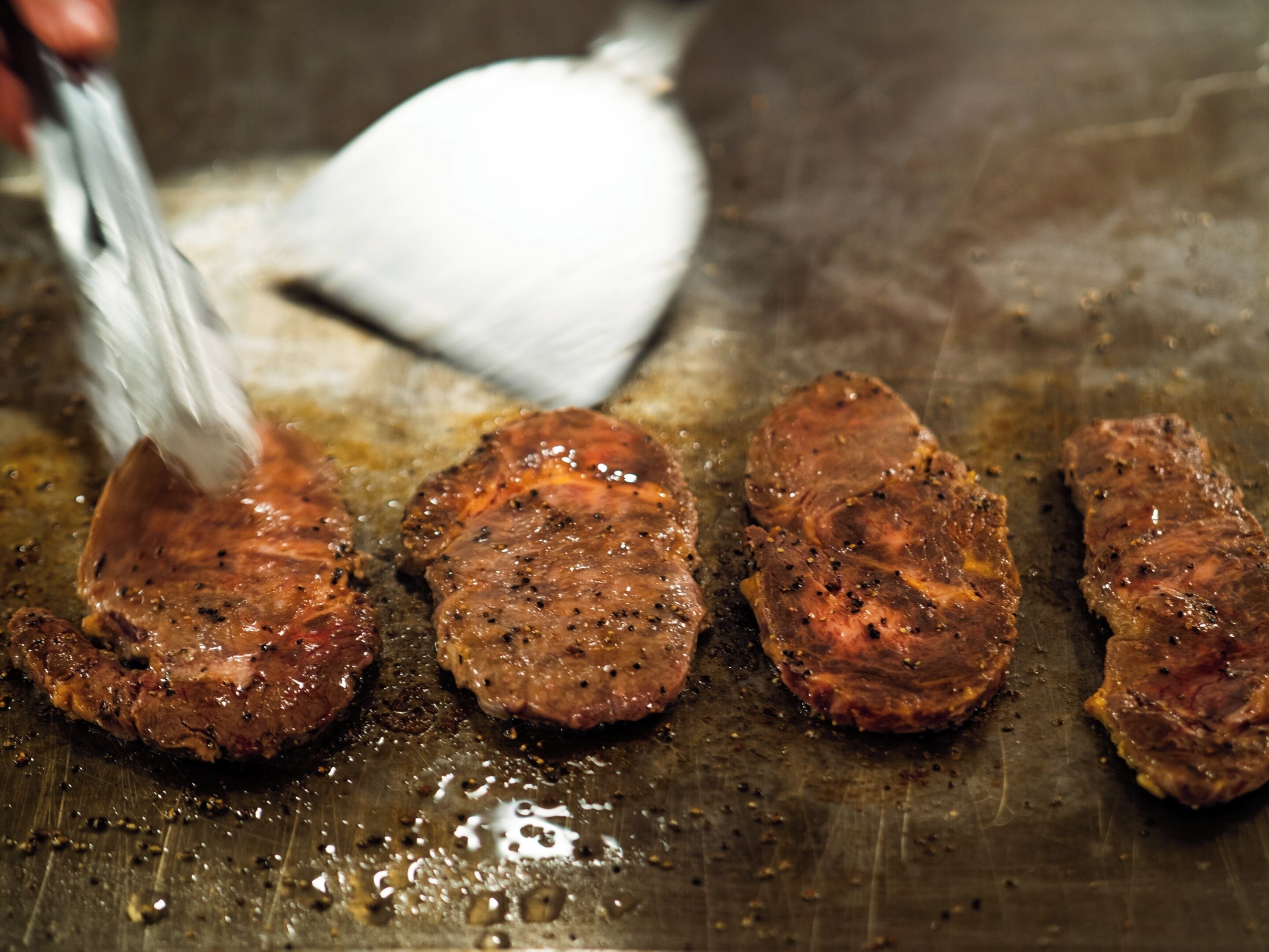 Gros plan sur quatre steaks en train de cuire sur un gril, une spatule à proximité.