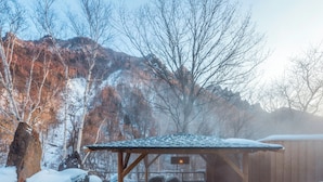 A wooden cabin with a snow-covered roof, surrounded by snow-covered rocks and trees, with a mountain in the background. - Sounkaku  (Kamikawa)