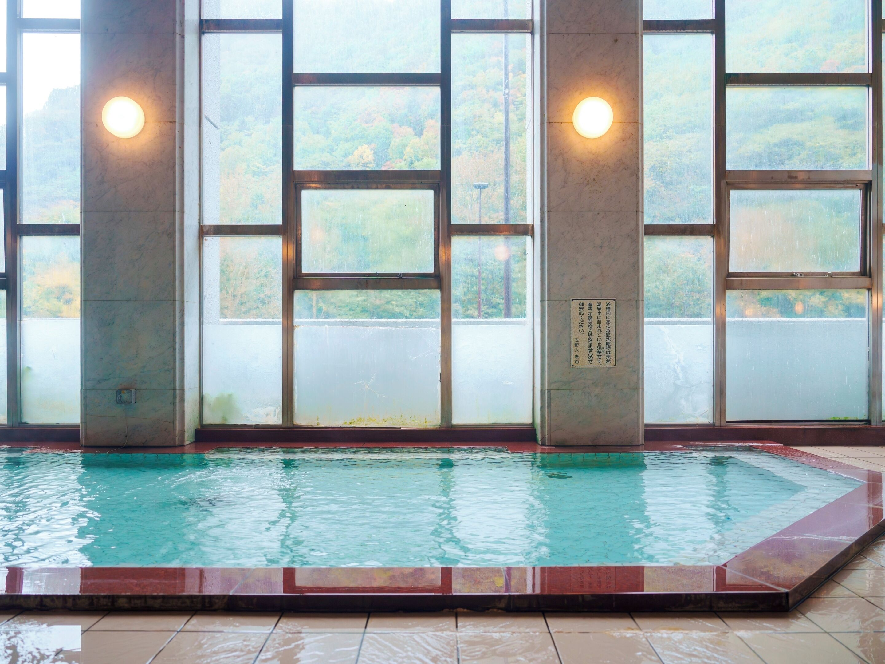 Una piscina con borde de azulejos, ventanas amplias con vidrio esmerilado y dos luces fijadas a la pared.