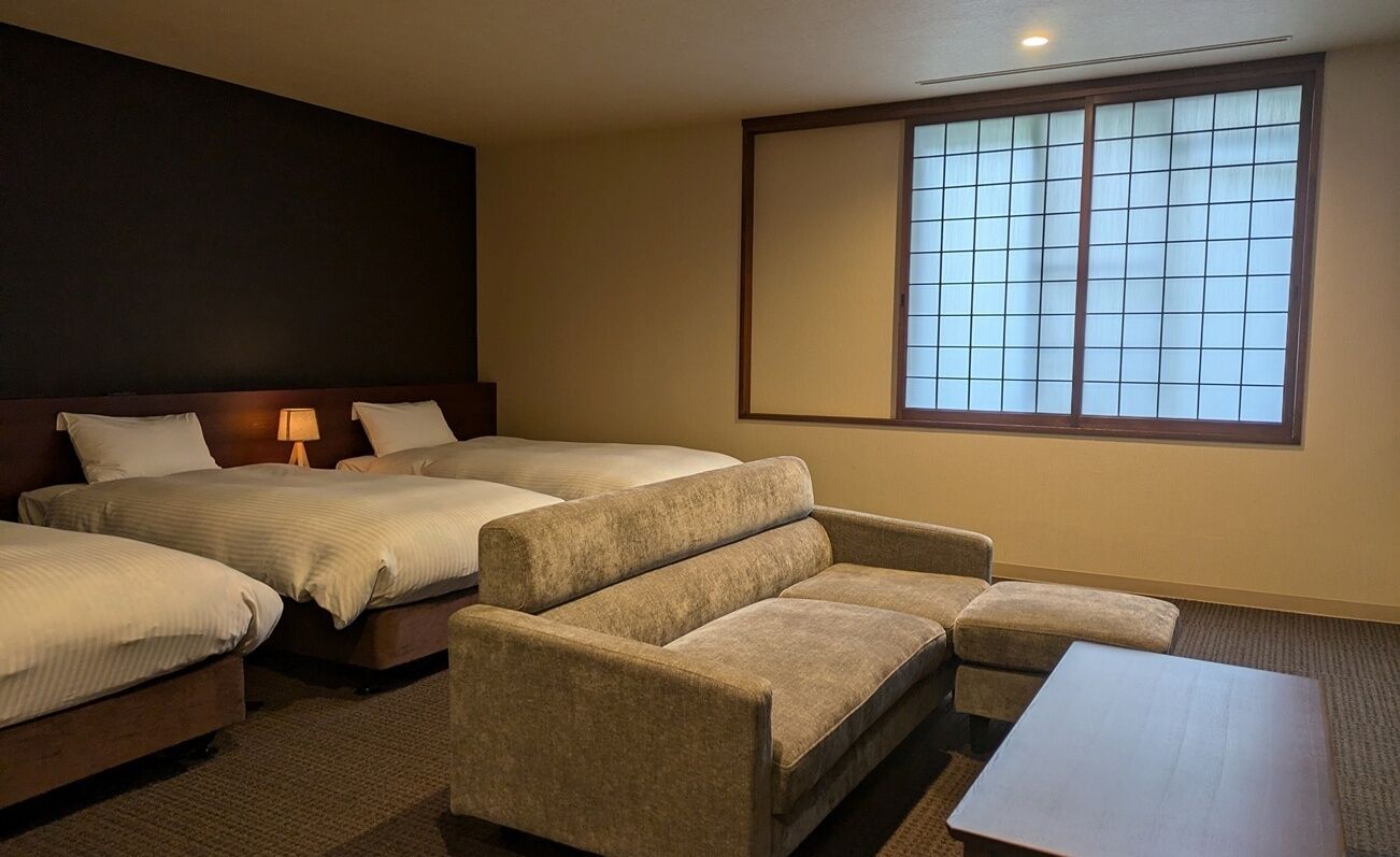 A hotel room with three beds, a sofa, a small table, and a large window with blinds.
