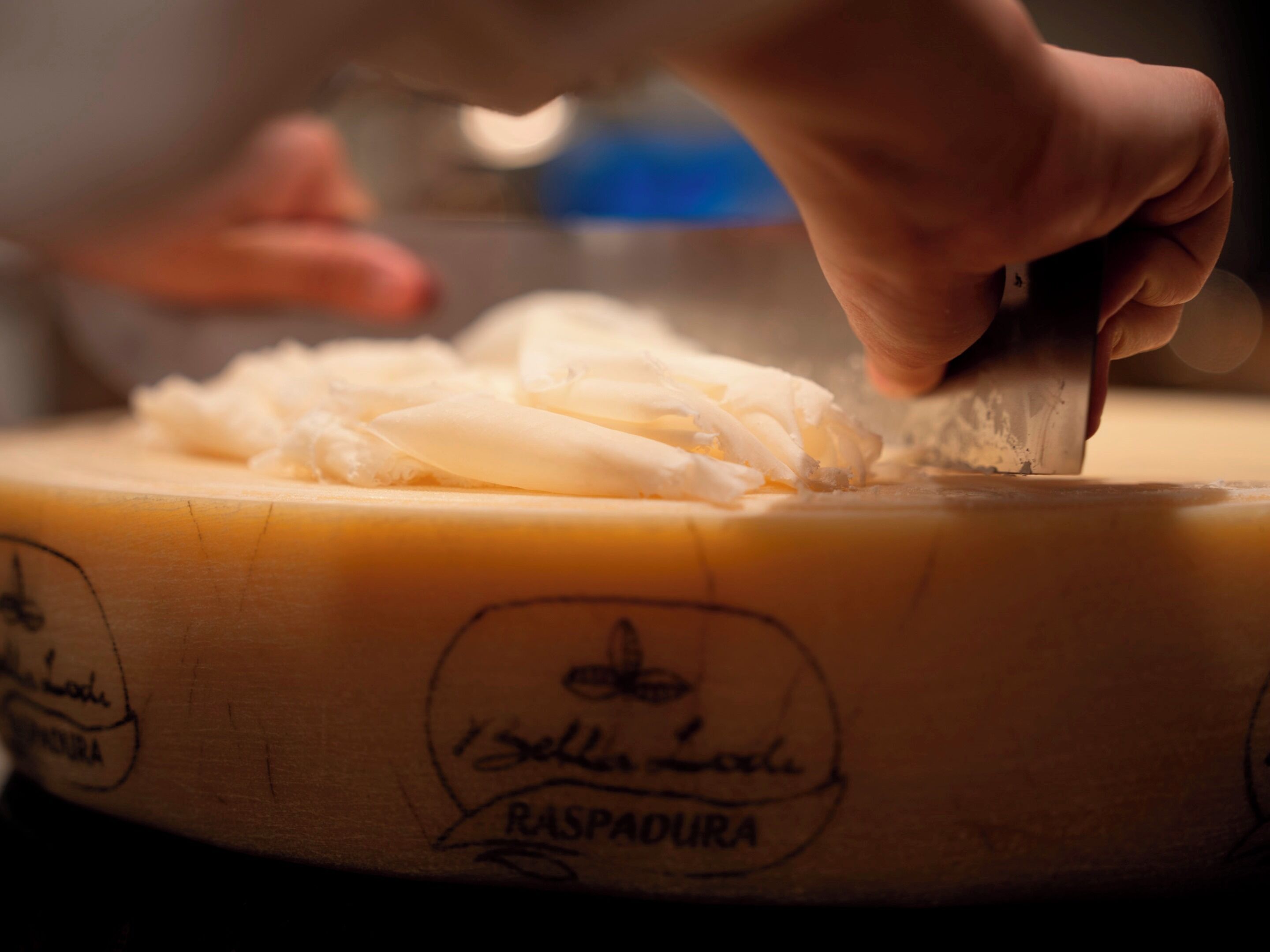 Una mano cortando un bloque grande de queso sobre una tabla de madera con la inscripción 'Isolina Lora RASPAUDURA'.