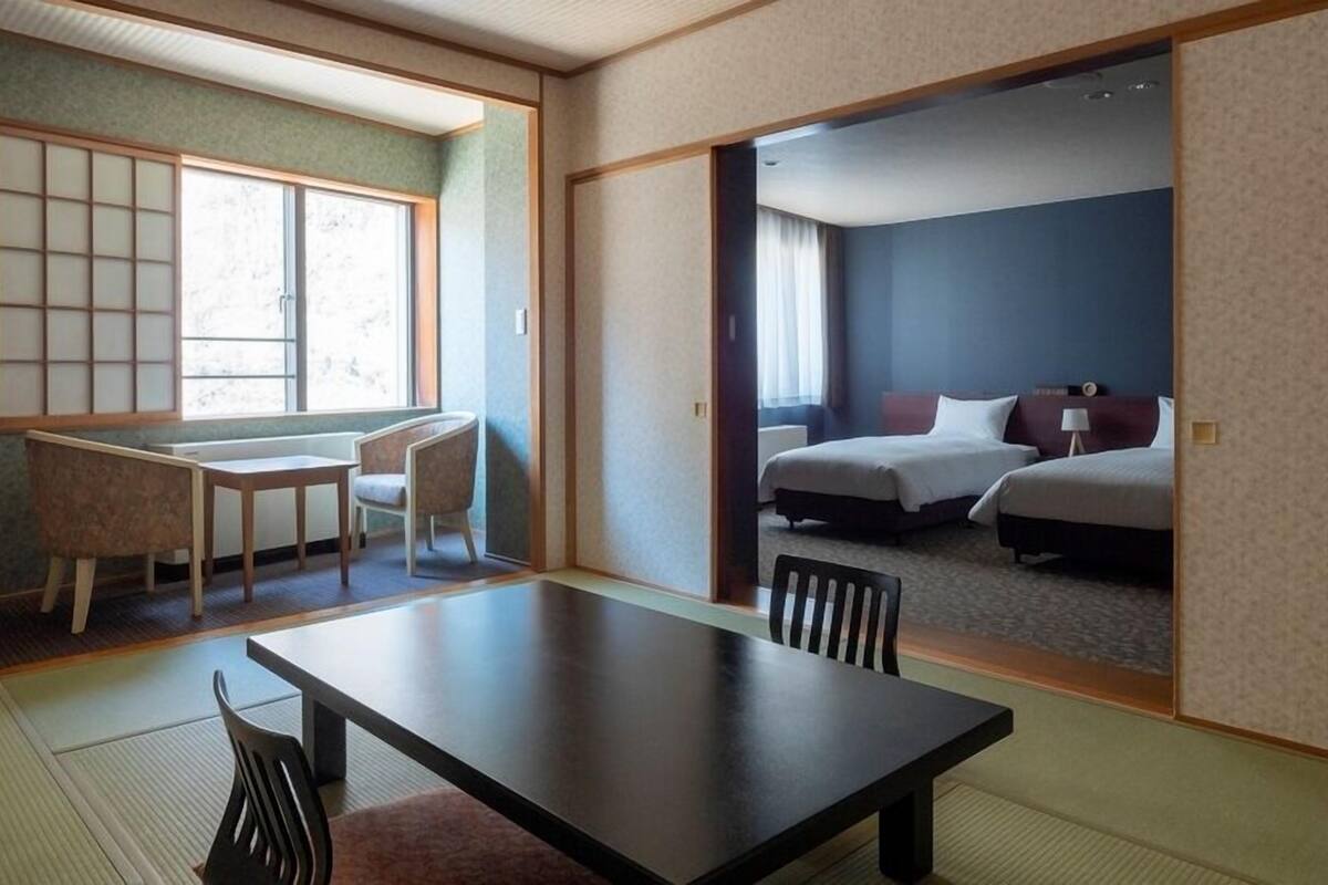 Una habitación de estilo japonés tradicional con suelo de tatami, una mesa baja y una puerta corredera que da a un dormitorio con dos camas.