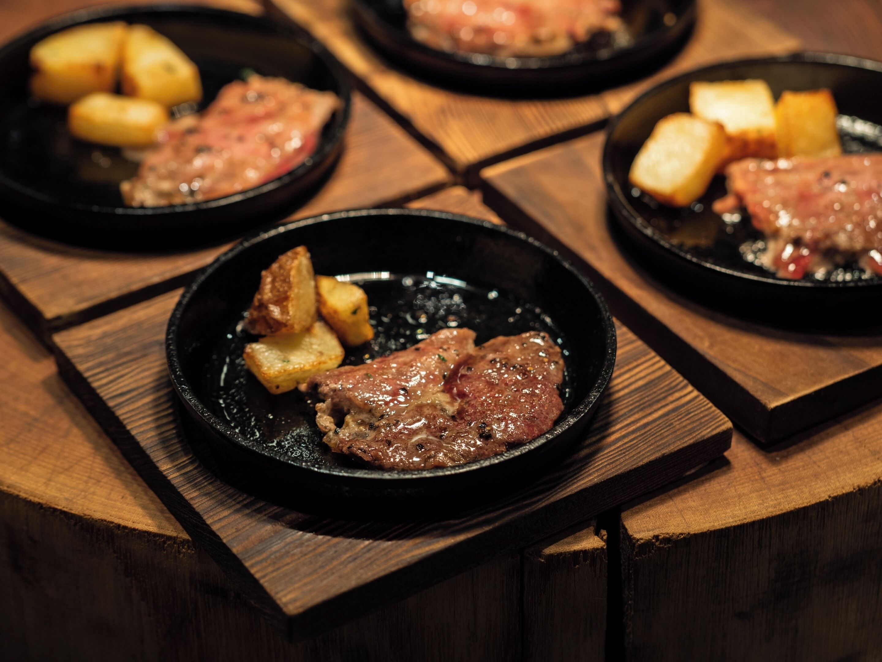 A wooden table with four black plates containing meat and potatoes.
