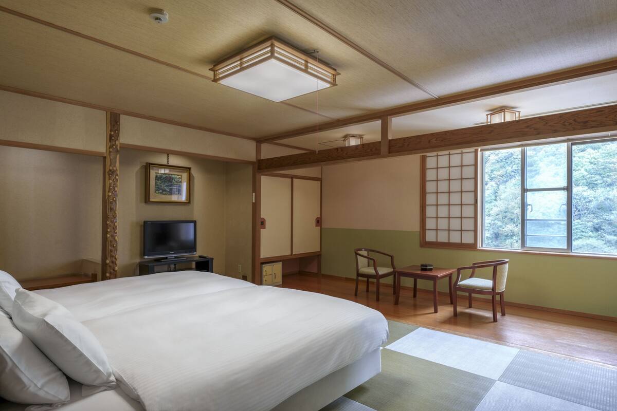 Habitación de hotel con cama, televisión, escritorio, sillas y ventana con vistas a árboles.