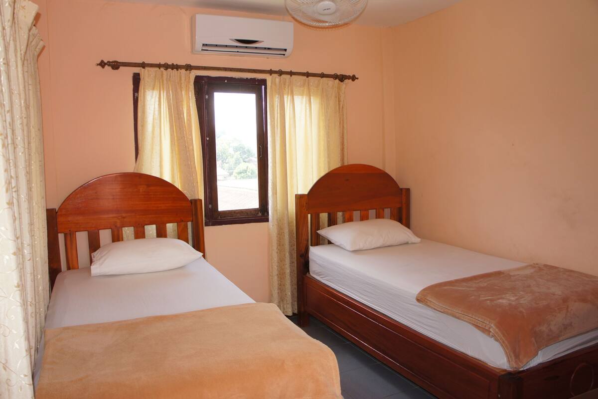 A room with two wooden beds, beige walls, a window with curtains, and an air conditioning unit.