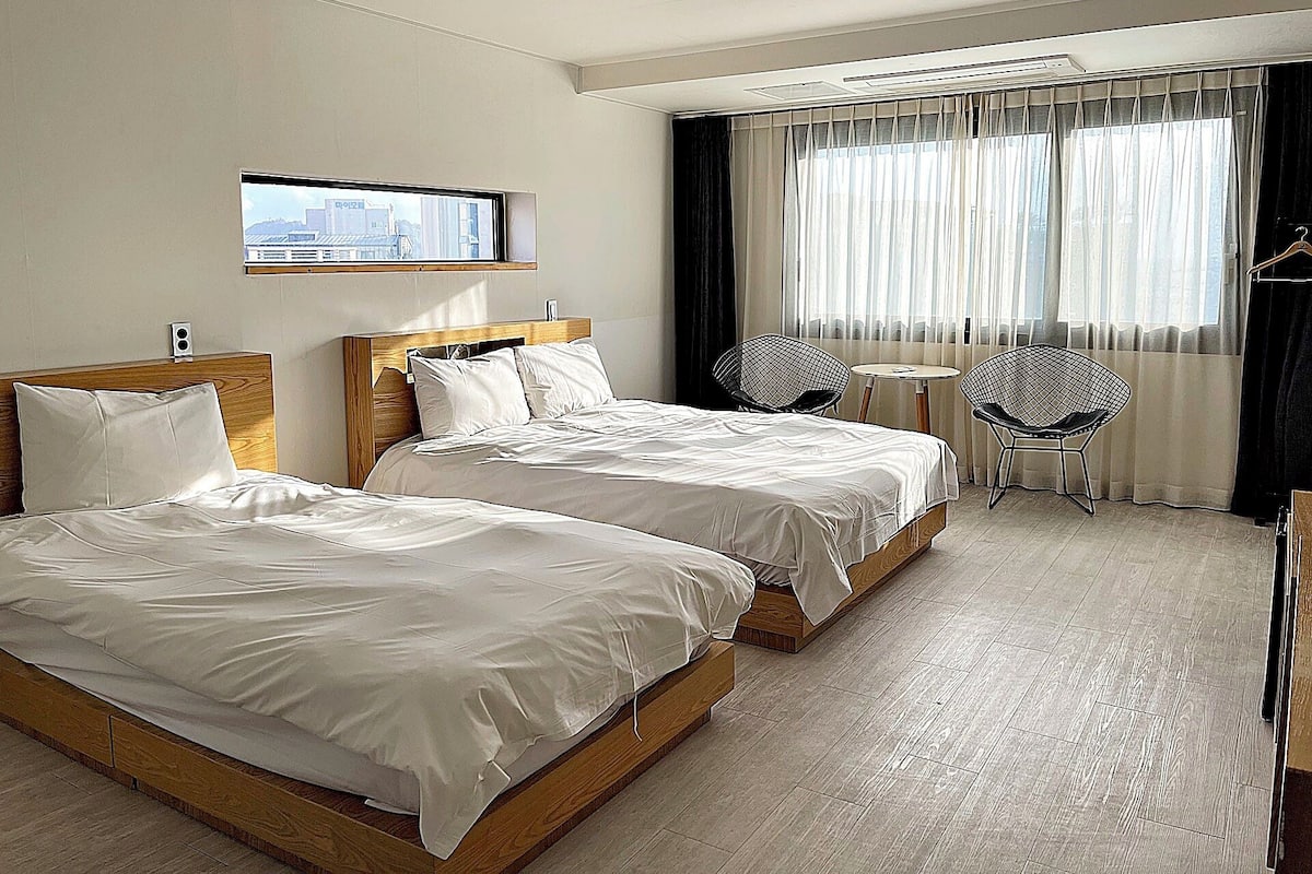 Habitación de hotel con dos camas, una mesita, dos sillas, una ventana con cortinas y vistas a la ciudad.