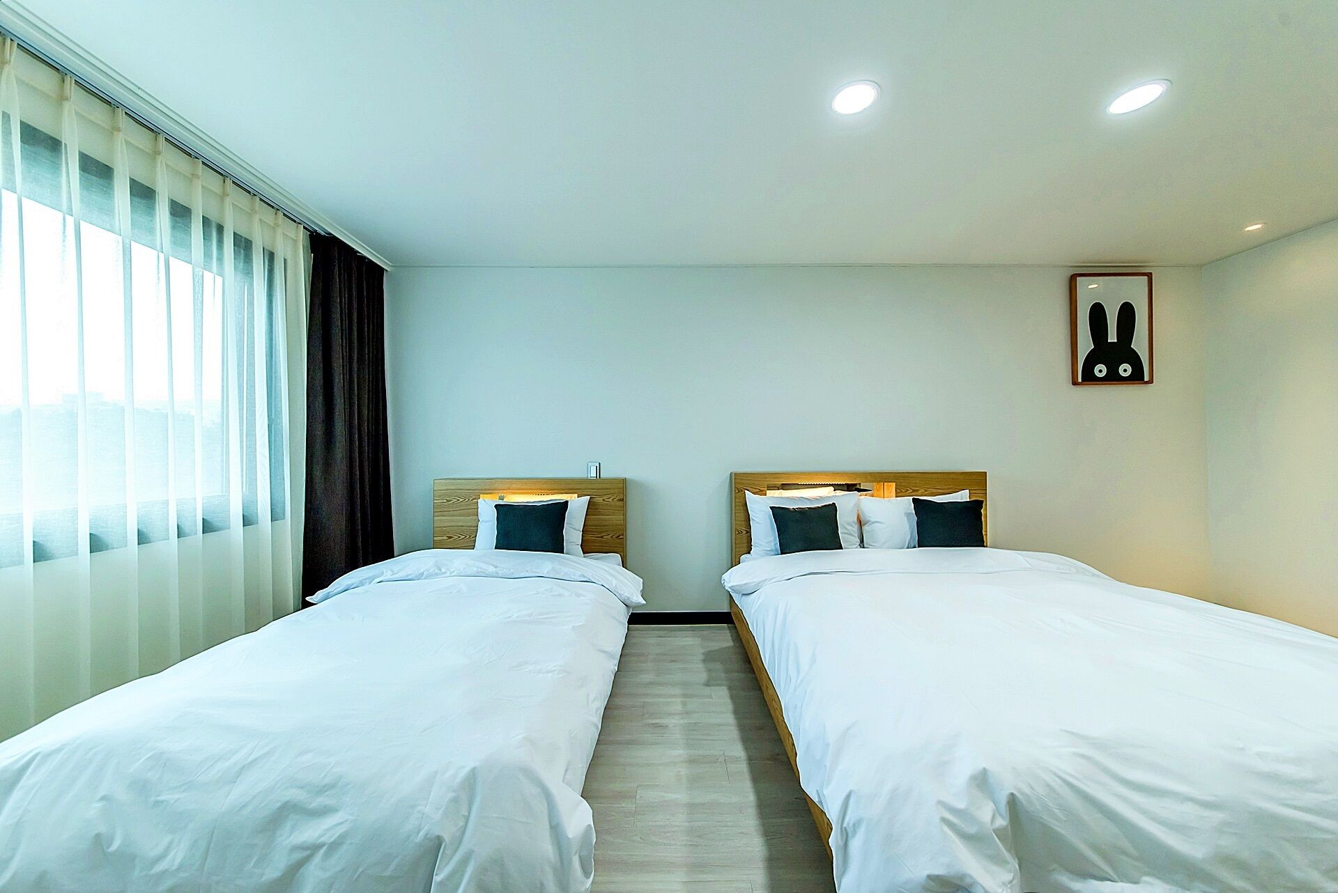 A hotel room with two single beds, a window with curtains, a wooden headboard, a small table, and a wall-mounted picture.