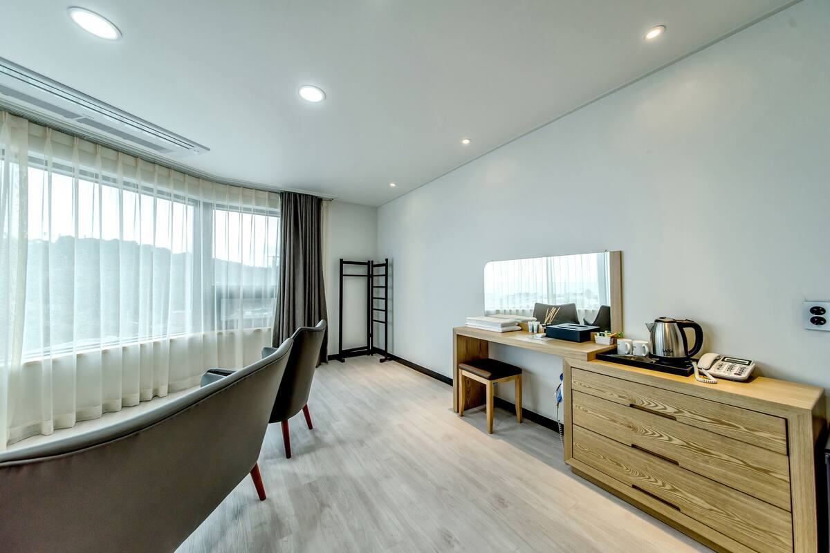 Una habitación de hotel moderna con un ventanal grande, un escritorio con computadora, una silla, un taburete pequeño y un aparador de madera con cajones.