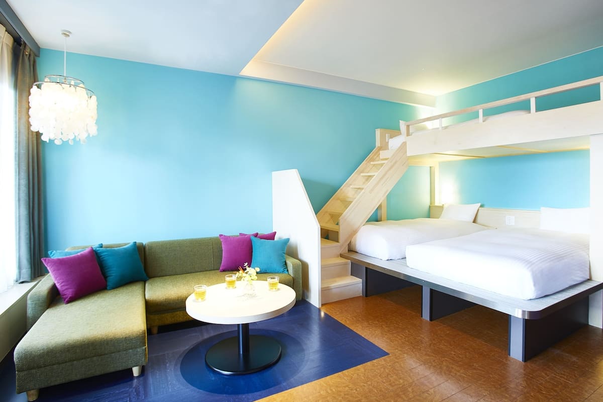 Une chambre d’hôtel avec un lit superposé, un canapé agrémenté de coussins colorés, une table ronde avec des boissons et un escalier.