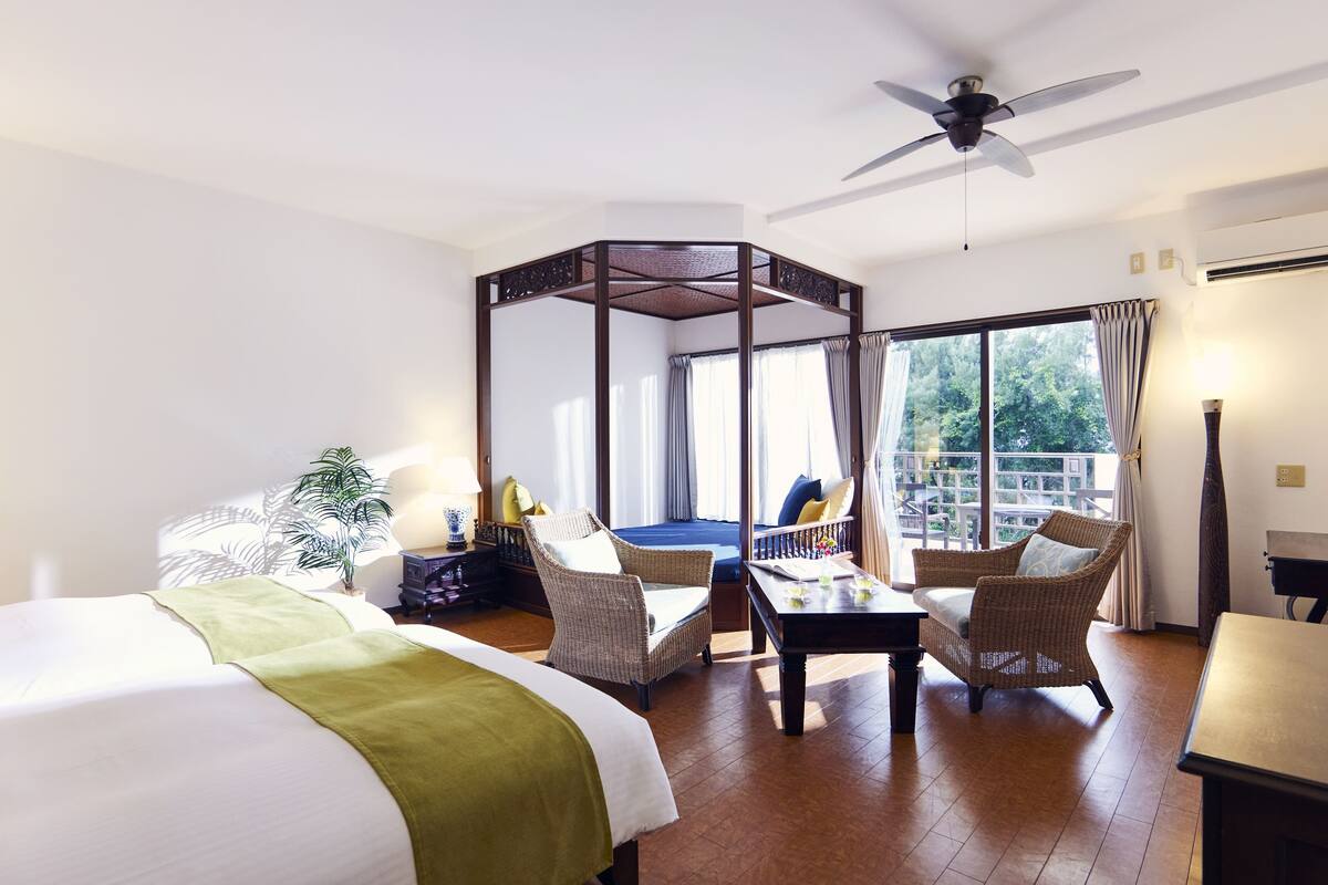 Une chambre d’hôtel avec un grand lit, deux fauteuils, une petite table, un ventilateur de plafond et un balcon donnant sur un paysage verdoyant.