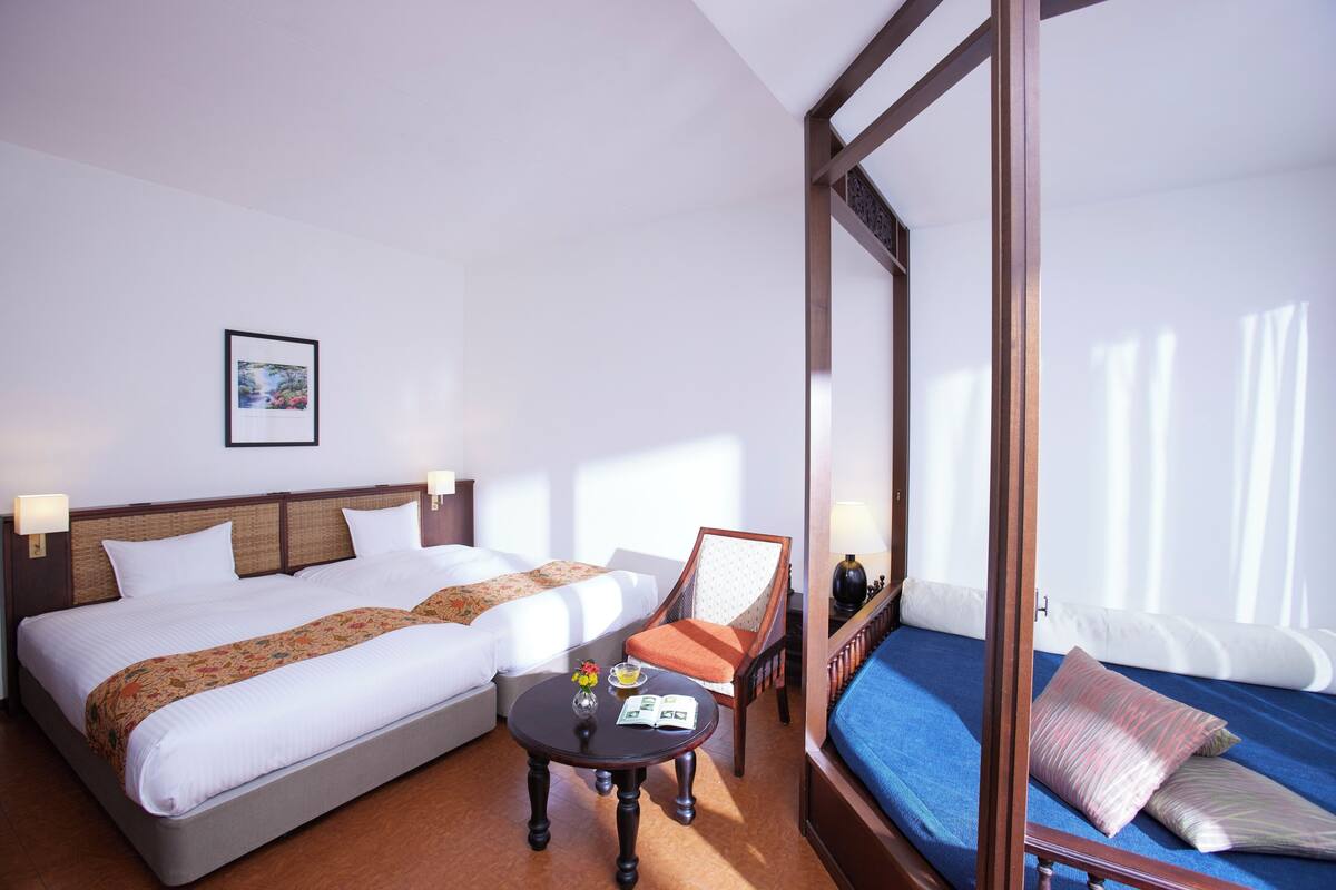 Une chambre d’hôtel avec un grand lit, un lit plus petit, une chaise, une table avec un vase et un tableau au mur.