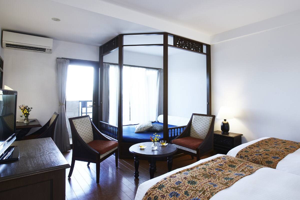 Une chambre d’hôtel avec un lit à baldaquin, deux fauteuils, une petite table et un bureau avec un ordinateur.