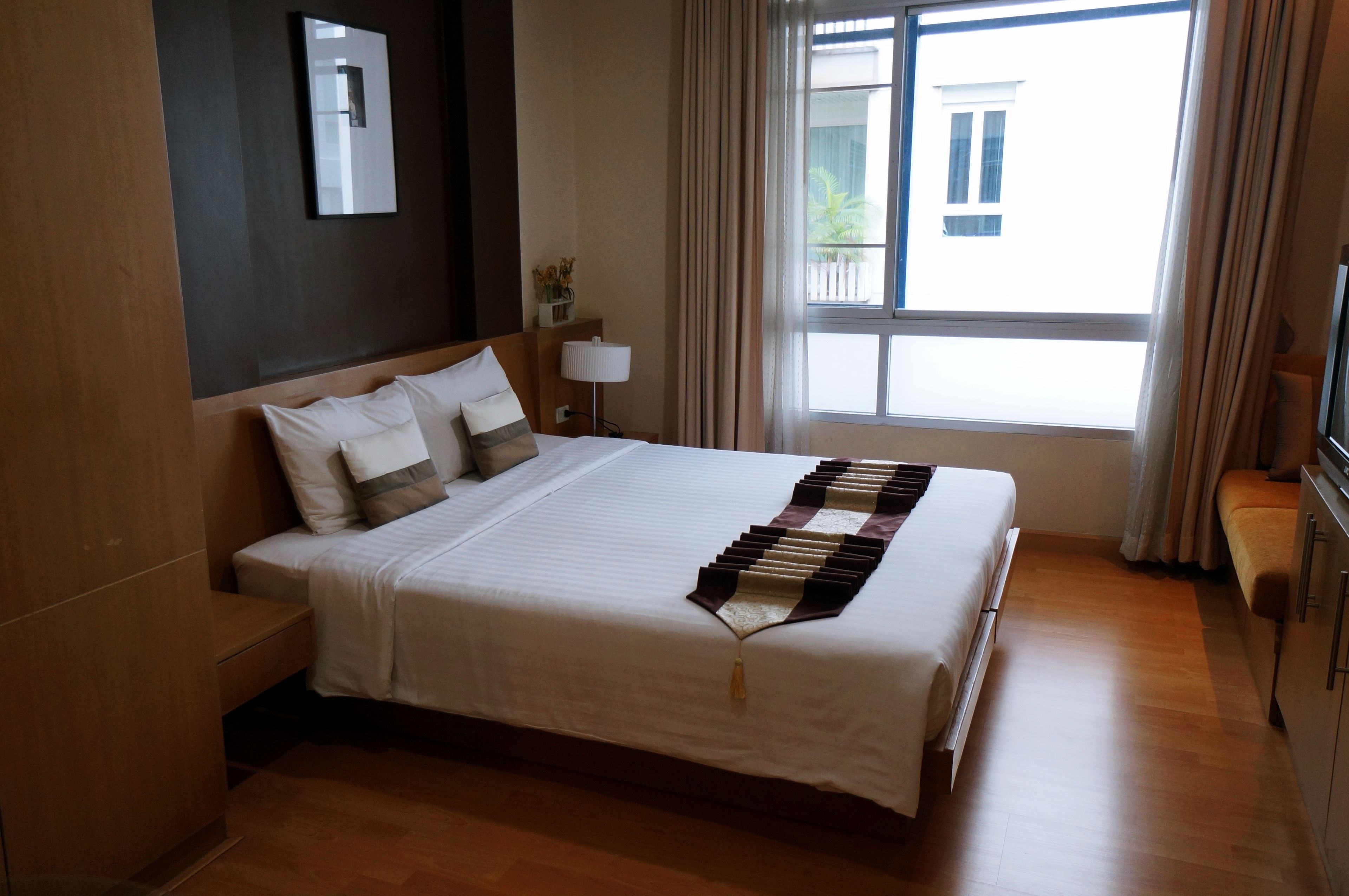 A hotel room with a large bed, a bedside table, a lamp, a TV, and a window with curtains.