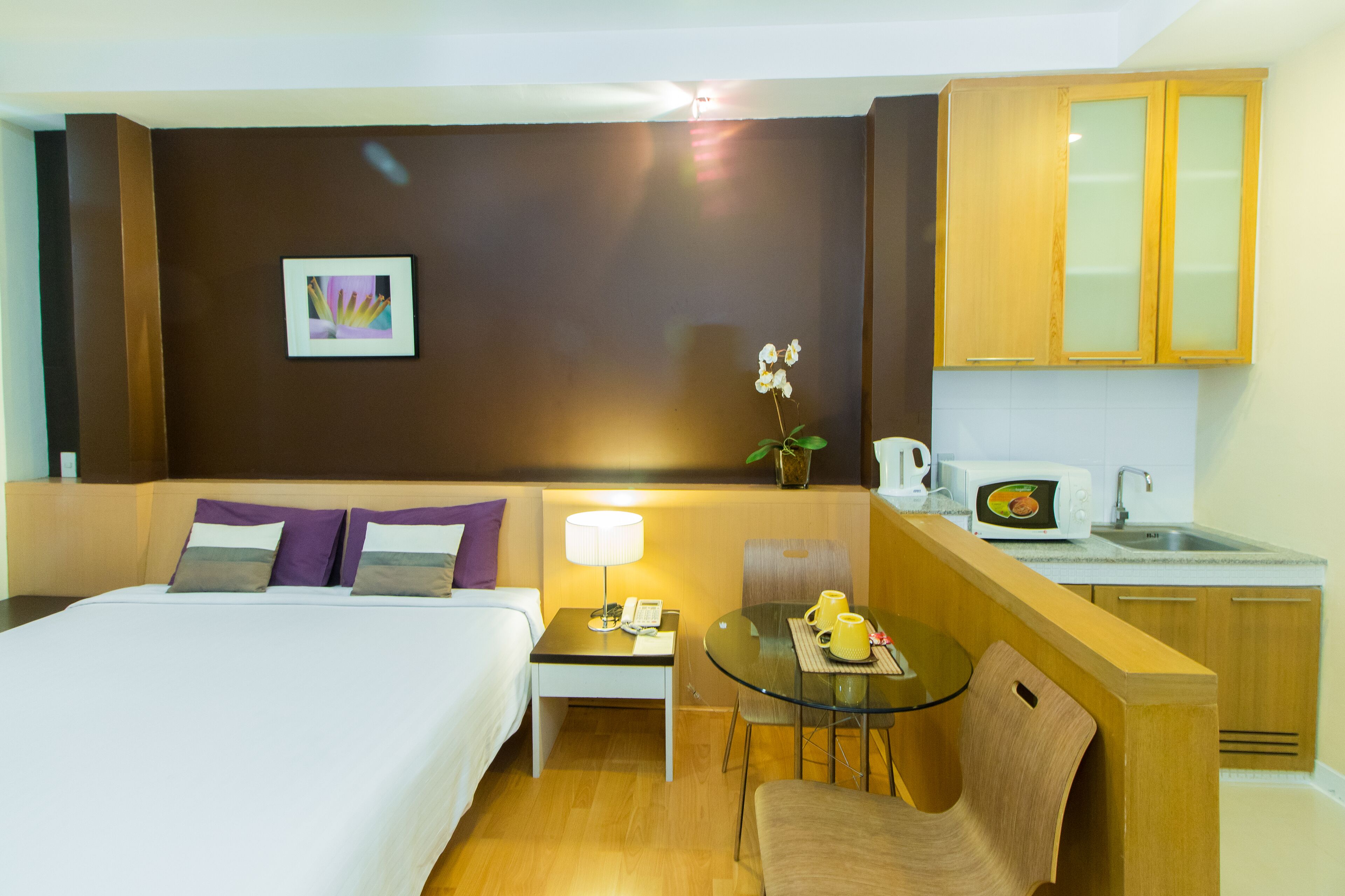 A hotel room with a bed, bedside table, lamp, chair, small table, and a kitchenette area.
