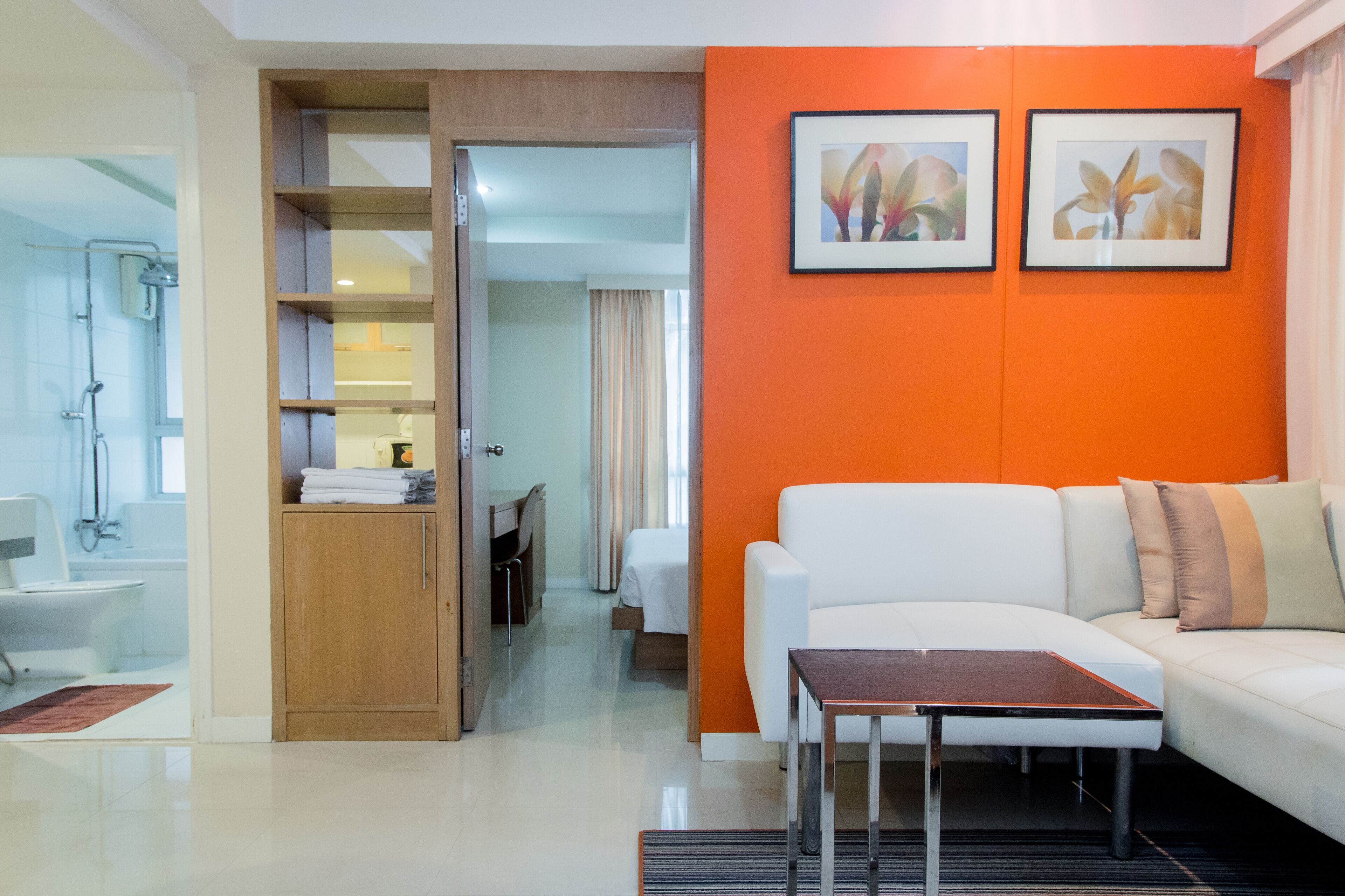 A modern hotel room with a white sofa, a small table, a wooden wardrobe, and an orange accent wall.