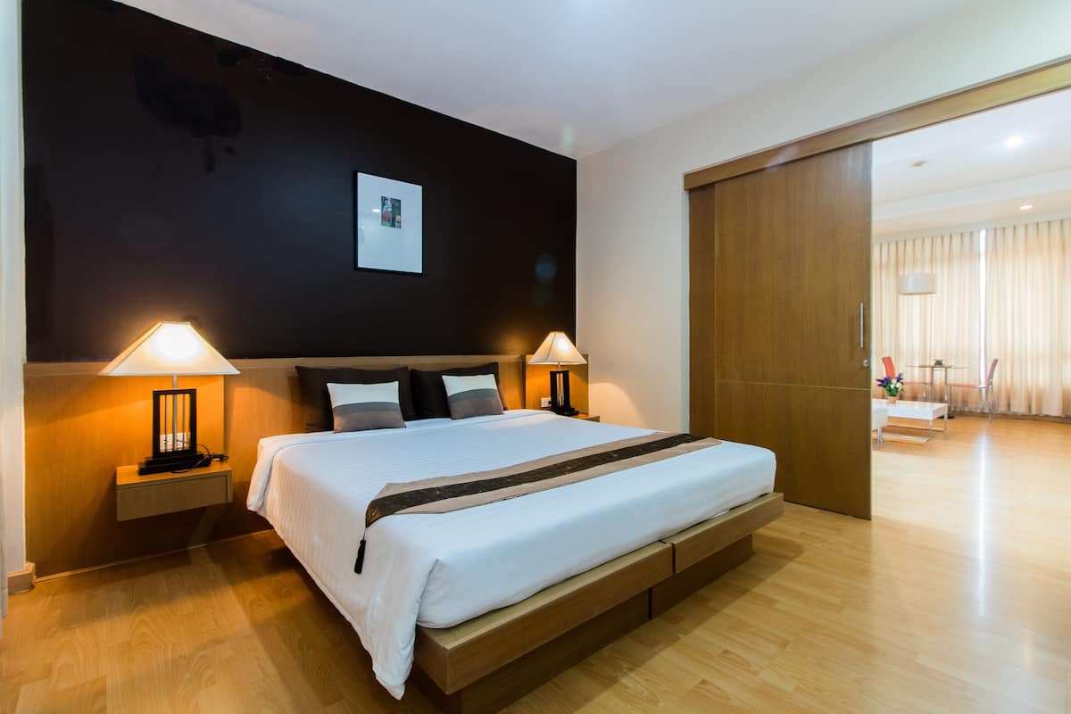 Moderni hotellihuone, jossa on suuri sänky, kaksi yöpöytävalaisinta, puulattia ja tumma seinä, jossa on kehystetty taulu.