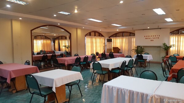 A banquet hall with multiple tables and chairs, a stage, and a banner reading "NAGUETE ROYAL SUITE INN".
