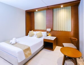 A hotel room with a large bed, a bedside table with a lamp, a chair, and a window with blinds.