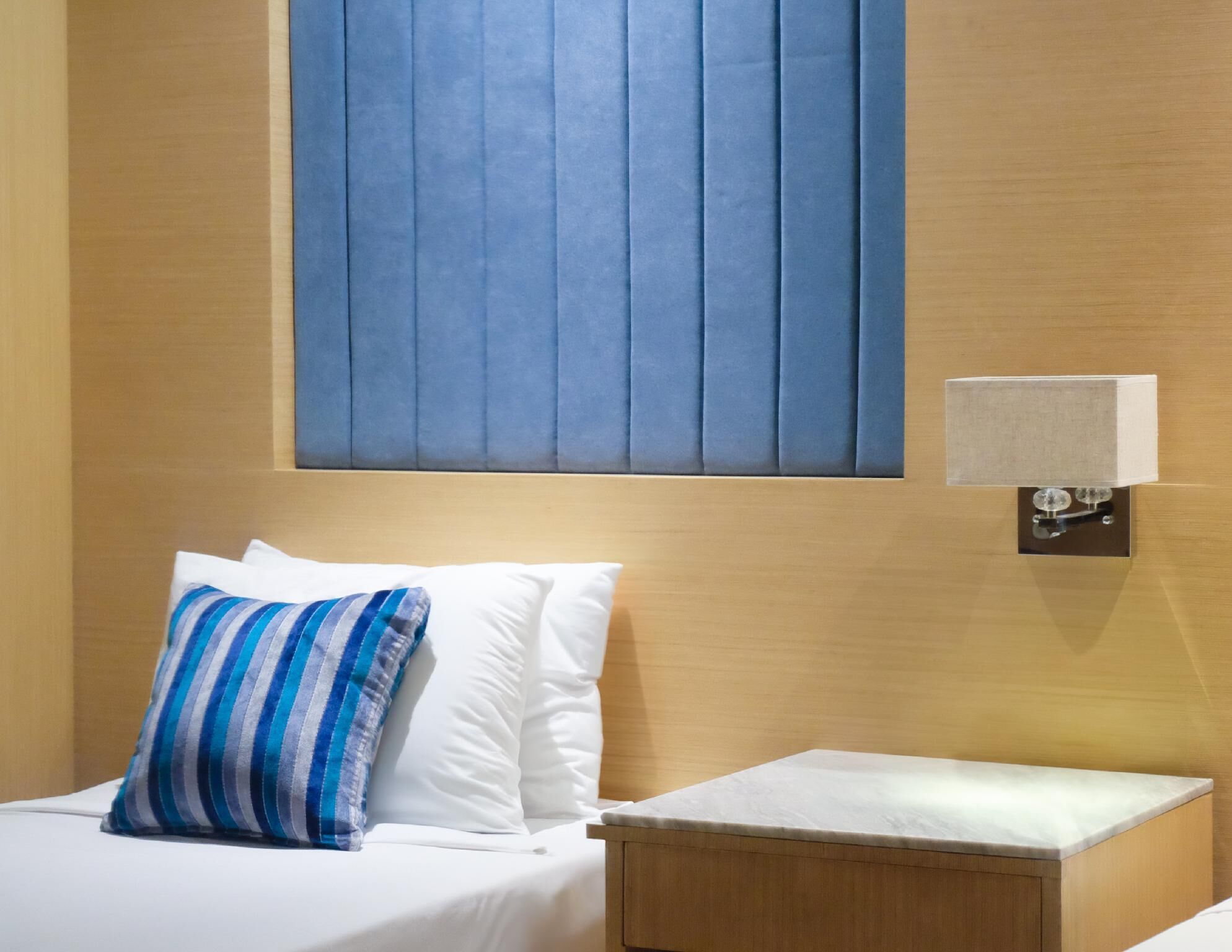 A hotel room with a bed, a bedside table, a wall-mounted lamp, and a window with vertical blinds.