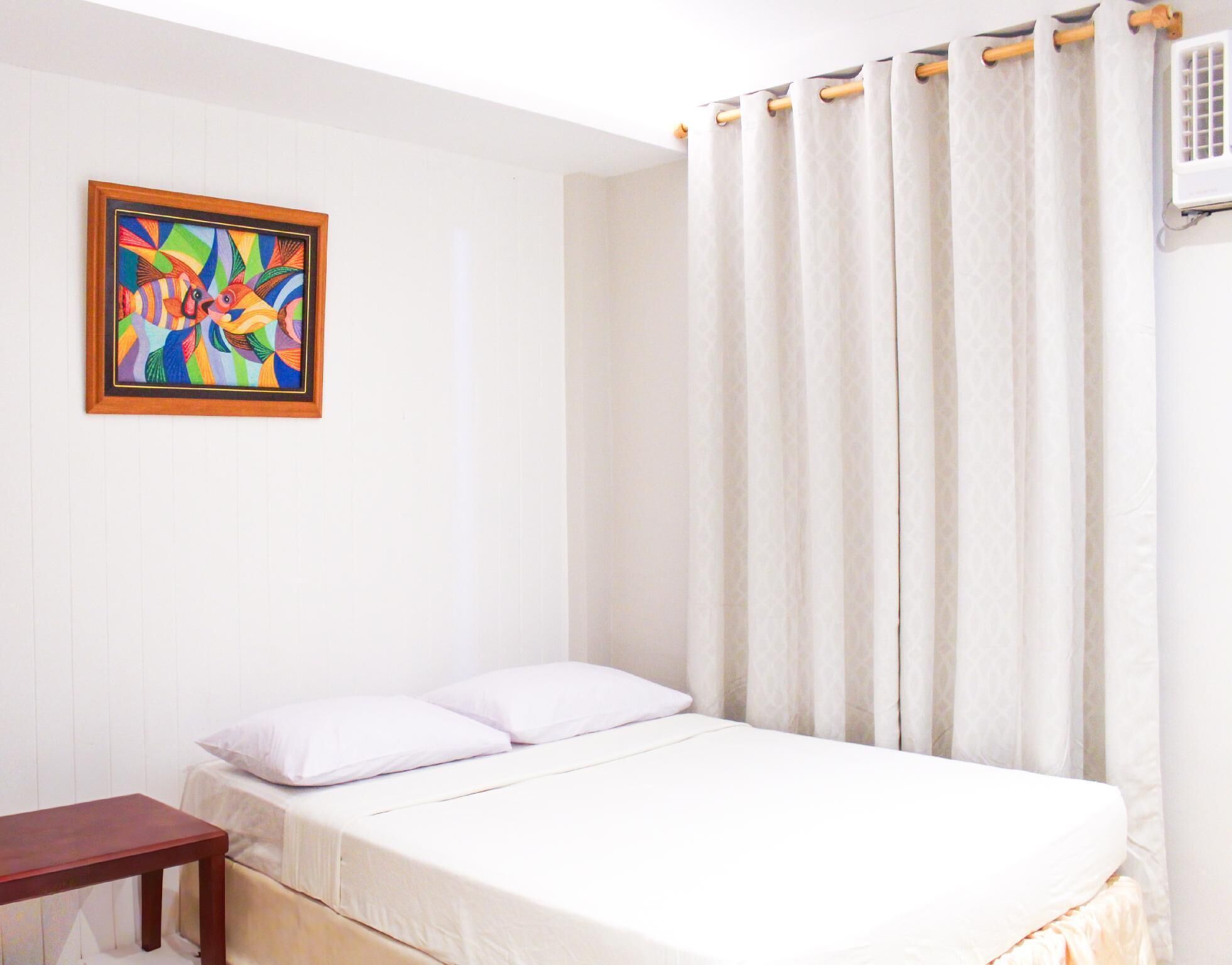 A single bed with white bedding, a wooden nightstand, a colorful abstract painting on the wall, and a window with white curtains.