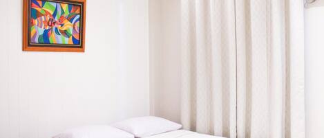 A single bed with white bedding, a wooden nightstand, a colorful abstract painting on the wall, and a window with white curtains.