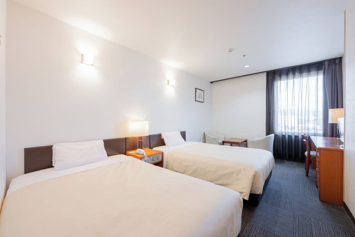 Une chambre d’hôtel avec deux lits, un bureau, une chaise et une fenêtre avec des rideaux.