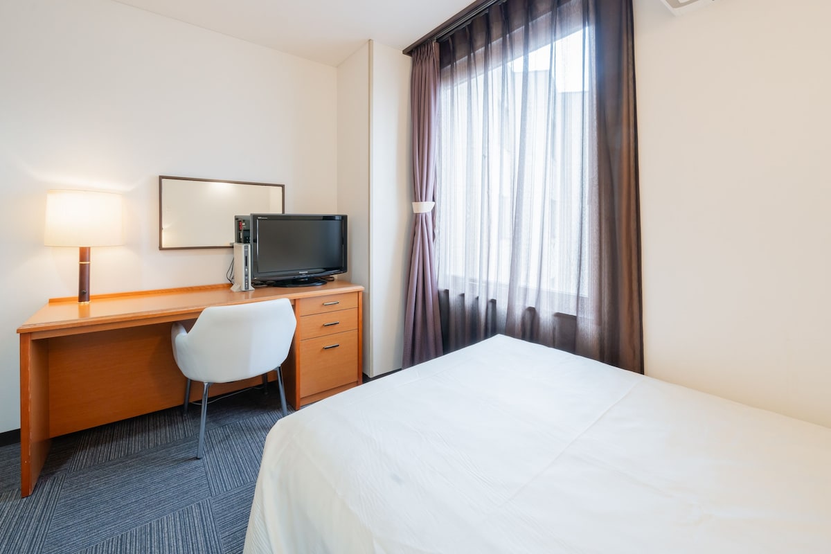 Habitación de hotel con cama, escritorio con televisor, una silla y ventana con cortinas.