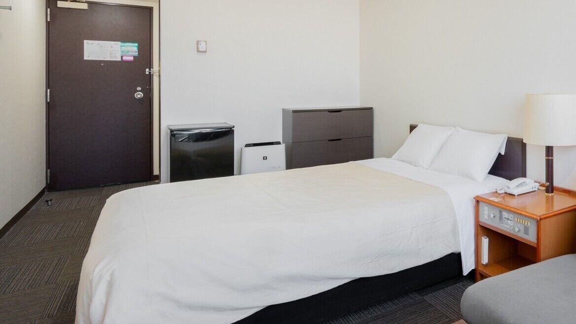 A hotel room with a bed, a desk, a chair, a lamp, a trash bin, and a filing cabinet.
