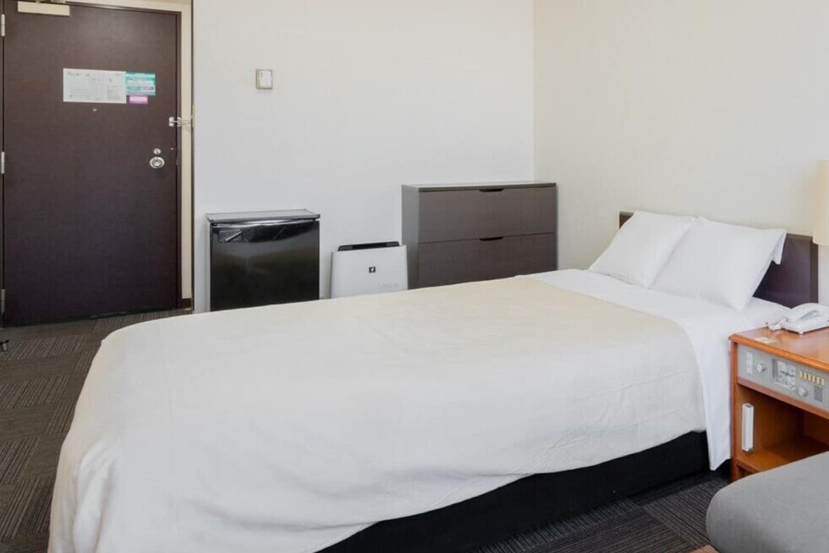 Quarto de hotel com cama, escrivaninha, cadeira, luminária, lixeira e armário para documentos.
