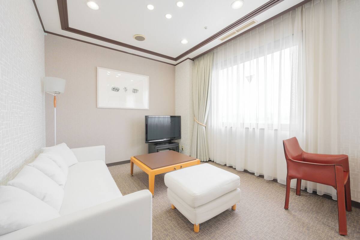 Une chambre d’hôtel avec un canapé blanc, une table basse en bois, un fauteuil rouge, une télévision et une grande fenêtre avec des rideaux.
