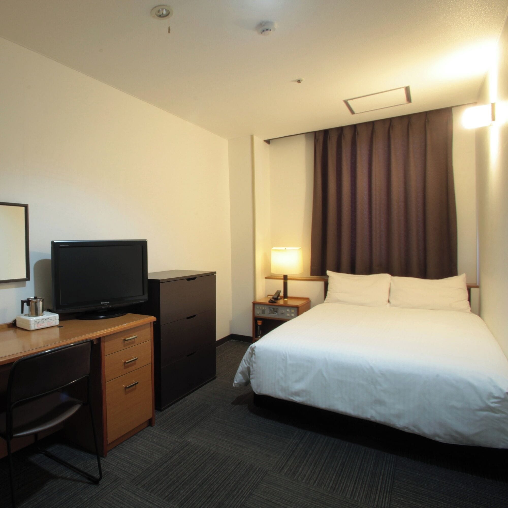 Una habitación de hotel con cama, escritorio con televisor, cómoda, mesita de noche con lámpara y ventana con cortinas.