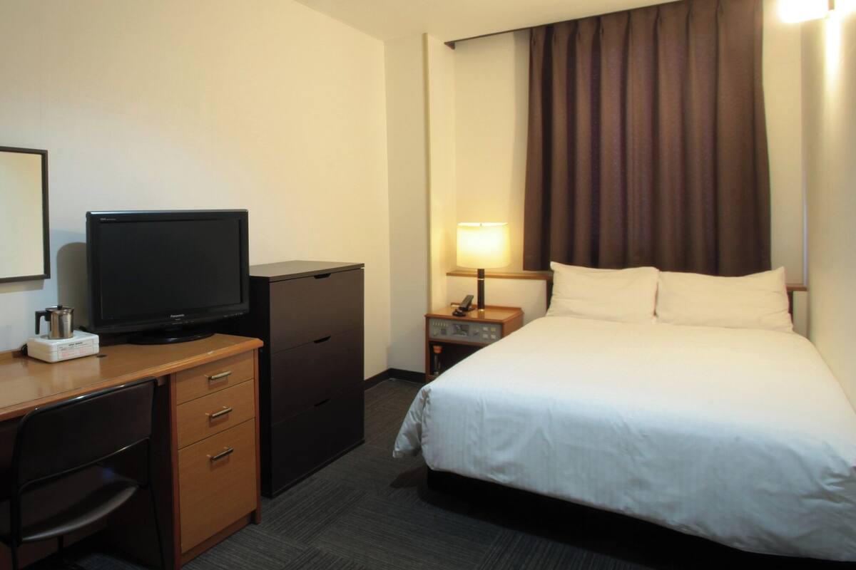 Une chambre d’hôtel comprenant un lit, un bureau avec une télévision, une commode, une table de chevet avec une lampe et une fenêtre avec des rideaux.