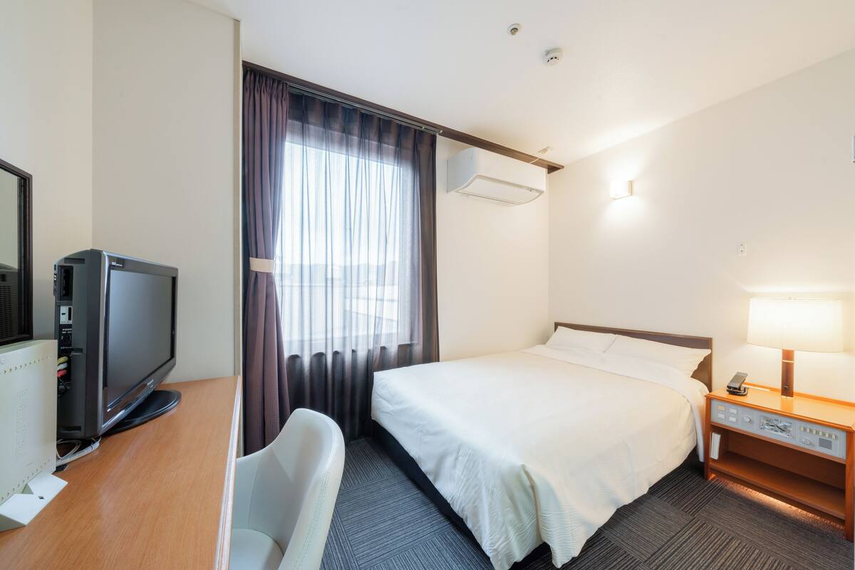 Une chambre d’hôtel comprenant un lit, un bureau avec une télévision, une chaise, une table de chevet avec une lampe et une fenêtre avec des rideaux.