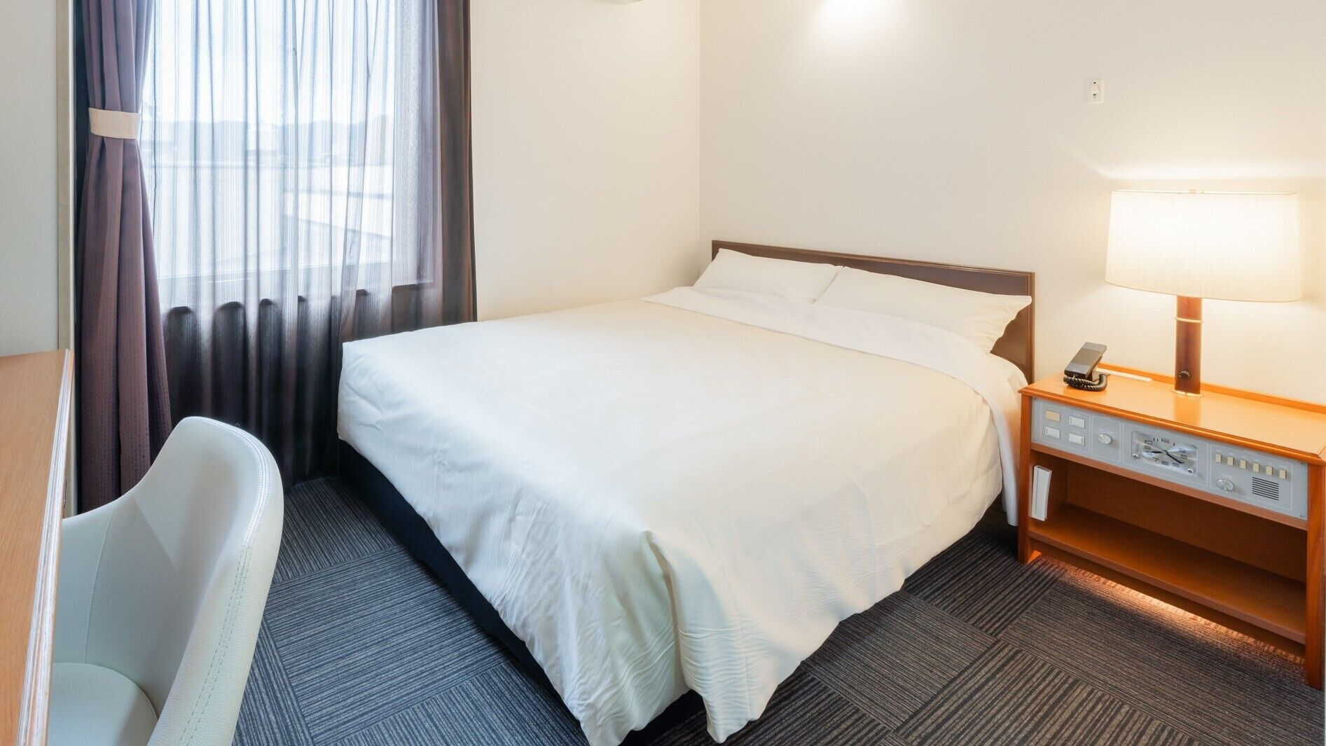 A hotel room with a bed, a chair, a bedside table with a lamp, and a clock.