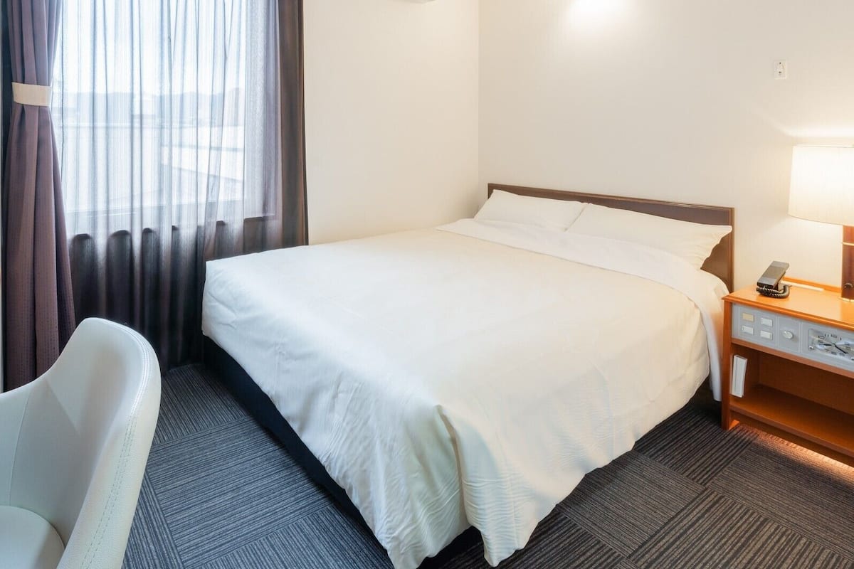 Quarto de hotel com uma cama, uma cadeira, mesa de cabeceira com abajur e um relógio.