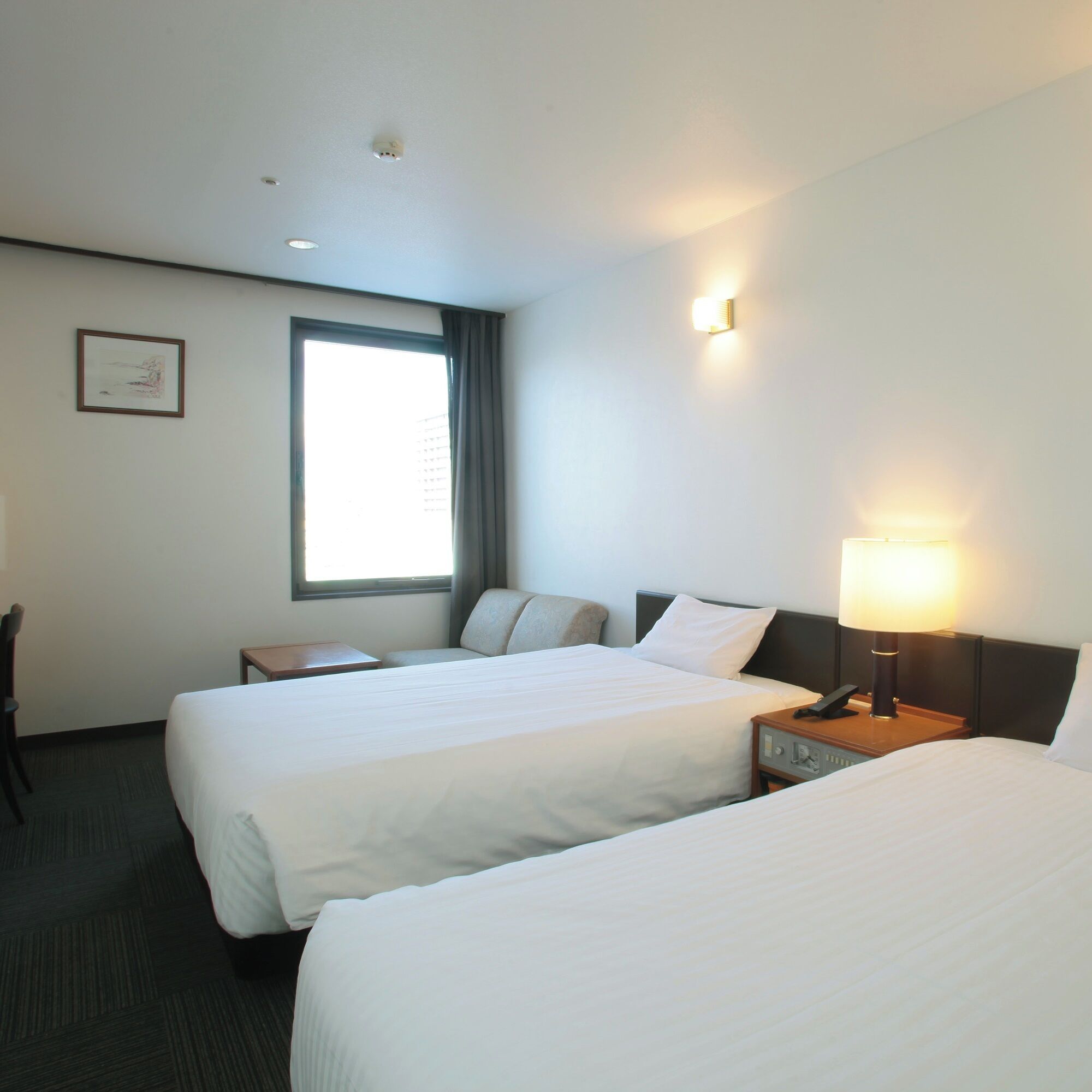 Habitación de hotel con dos camas individuales, un escritorio, una silla, una ventana con cortinas y una lámpara fijada a la pared.