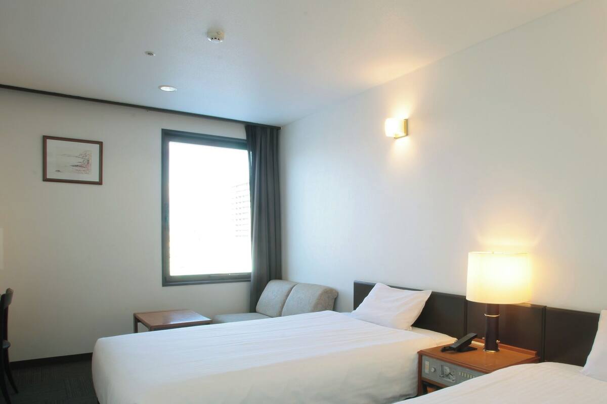 Une chambre d’hôtel avec deux lits simples, un bureau, une chaise, une fenêtre avec des rideaux et un luminaire fixé au mur.