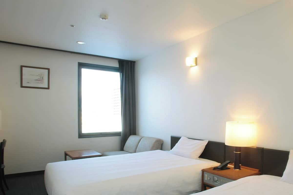 A hotel room with two single beds, a desk, a chair, a window with curtains, and a wall-mounted light fixture.