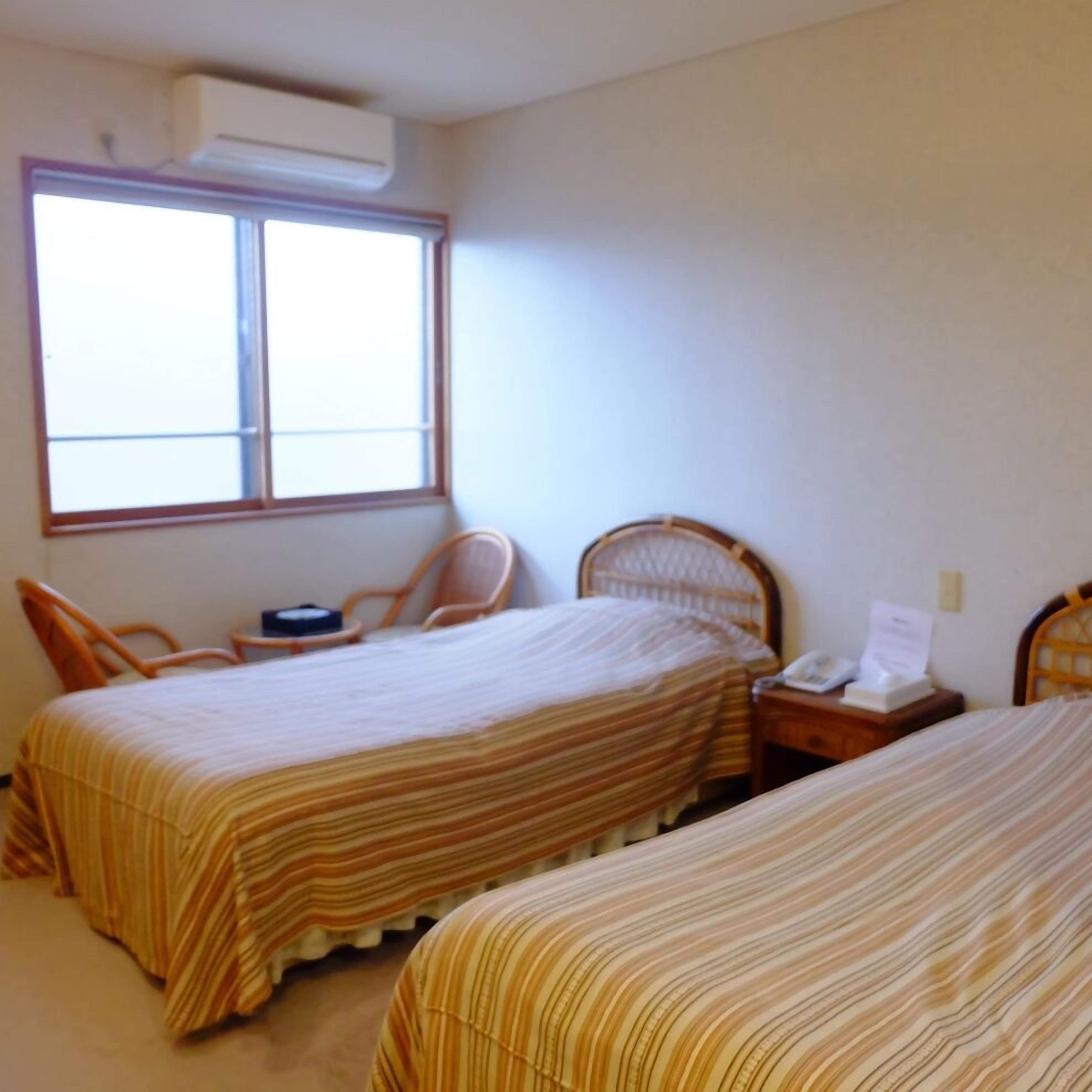 A room with two beds, a window, an air conditioning unit, and a chair.