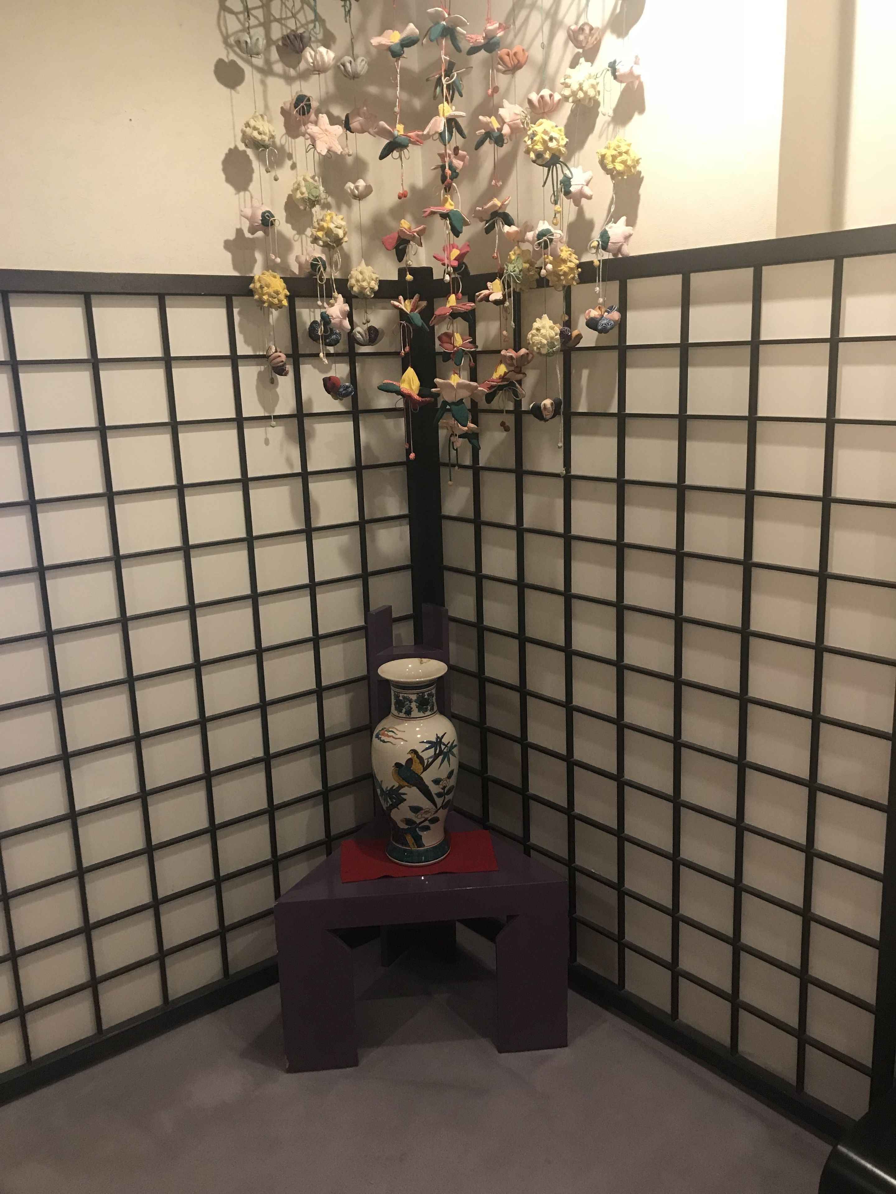 A decorative vase with a bird design on a pedestal in a corner with a grid-patterned wall.