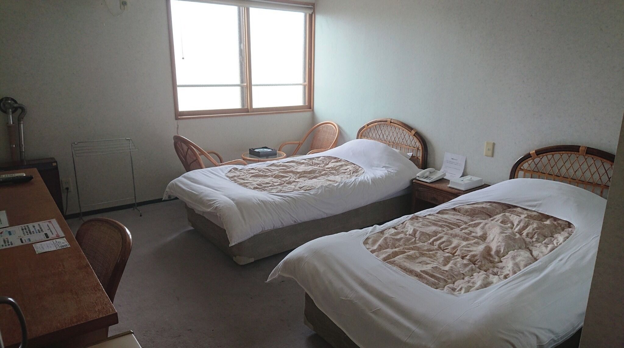 A hotel room with two single beds, a wooden desk, a chair, a radiator, and a window providing natural light.