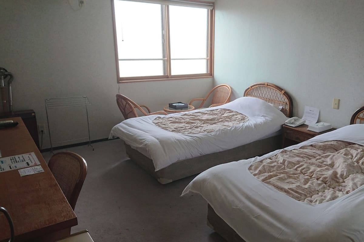 Une chambre d’hôtel avec deux lits simples, un bureau en bois, une chaise, un radiateur et une fenêtre laissant entrer la lumière naturelle.